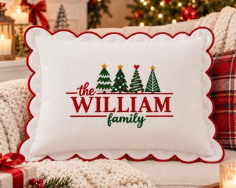 May include: A white rectangular throw pillow with a scalloped red edge. The center features embroidered Christmas trees and the red and green text "the WILLIAM family". The pillow is positioned on a couch with a red plaid cushion nearby.