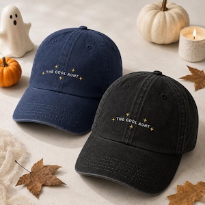 May include: A navy blue baseball cap and a black baseball cap, both featuring the embroidered text THE COOL AUNT in white capital letters, accented by small yellow four-pointed star icons on the front panels of each hat.