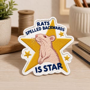 May include: An embroidered patch featuring a light pink rat centered on a large yellow star. The text RATS SPELLED BACKWARDS IS STAR is stitched in dark blue thread around the star. The patch has a white border and includes three small yellow stars.