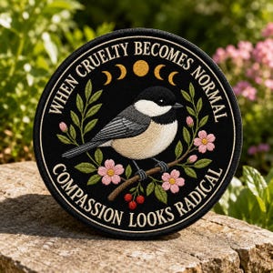 May include: A circular black embroidered patch featuring a chickadee perched on a branch with pink flowers and red berries. Above the bird are golden moon phases. The text reads: WHEN CRUELTY BECOMES NORMAL COMPASSION LOOKS RADICAL.
