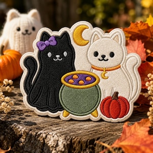 May include: An embroidered patch featuring a black cat with a purple bow and a white cat with an orange collar and crescent moon charm. Between them sits a green cauldron filled with purple liquid and orange bubbles, next to a small orange pumpkin.