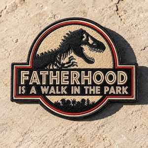 May include: Embroidered patch with a black dinosaur silhouette and the text "FATHERHOOD IS A WALK IN THE PARK" in white. The patch has a tan background with a red and black border, suitable for jackets or bags.
