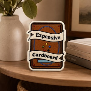 May include: A rectangular embroidered patch featuring a brown background with a textured leather-like appearance, a blue circular border, and six small colored dots. Two white banners with black text read Expensive and Cardboard.