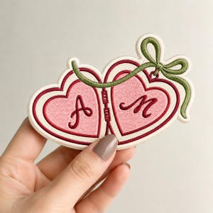May include: An embroidered patch featuring two connected heart shapes in shades of pink, outlined in dark red and cream. The left heart displays the letter A and the right heart displays the letter M in cursive script, with a green ribbon bow on top.