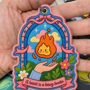 May include: Embroidered keychain with a blue and pink arched design. The keychain features a flame with a face, a hand, flowers, and the text "A heart is a heavy burden."