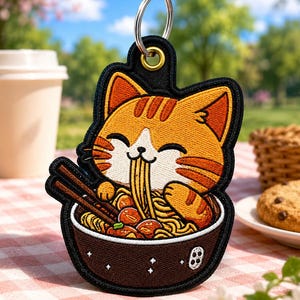May include: A keychain featuring an embroidered orange tabby cat enjoying ramen noodles. The cat is smiling and holding chopsticks. The keychain has a black border and a silver keyring. The background shows a picnic setting with a coffee cup and cookies.