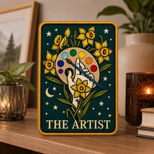 May include: A rectangular embroidered patch on a dark teal background featuring a hand holding a paintbrush over a paint palette, surrounded by yellow daffodils, stars, and a crescent moon, with the text THE ARTIST printed at the bottom.
