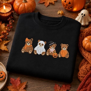May include: A black crewneck sweatshirt featuring an embroidered design of four brown teddy bears. The first bear wears an orange hat and scarf, the second is dressed as a ghost, the third wears a witch hat and pink bow, and the fourth holds a pumpkin.