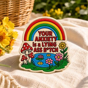 May include: A cream-colored embroidered patch featuring a rainbow arch over the text 'YOUR ANXIETY is a LYING ASS B*TCH'. The design includes red and white spotted mushrooms, a blue pond with pink and yellow flowers, and green grass.
