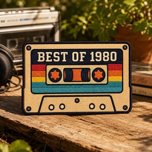 May include: A rectangular embroidered patch shaped like a cassette tape with a tan border and dark blue trim. The center features the text BEST OF 1980 in white letters above horizontal stripes of red, orange, yellow, and teal. It has two orange spools.