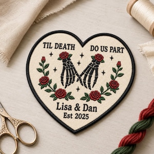 May include: A heart-shaped embroidered patch with a cream background and black border. It features two black skeleton hands holding each other, surrounded by red roses and green leaves. Text reads: TIL DEATH DO US PART, Lisa & Dan, Est 2025.