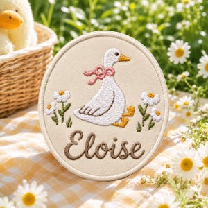 May include: A circular beige patch featuring an embroidered white goose with a pink bow around its neck and yellow feet. The goose is flanked by embroidered white daisies with green stems. The name Eloise is stitched in brown cursive text below.