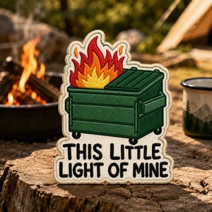 May include: An embroidered patch featuring a green dumpster on fire with bright red, orange, and yellow flames. Below the dumpster, the text reads THIS LITTLE LIGHT OF MINE in bold black letters. The patch has a cream-colored border.