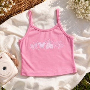 May include: A pink tank top featuring a white embroidered graphic across the chest. The design includes a bow, a Mickey Mouse head silhouette, a castle, and two hearts, all surrounded by small star accents. The top has thin shoulder straps.
