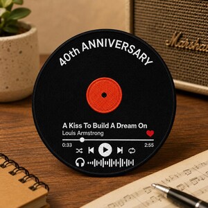 May include: A circular black embroidered patch shaped like a vinyl record. It features white text reading 40th ANNIVERSARY, A Kiss To Build A Dream On, and Louis Armstrong. A red center circle, a heart icon, and music player controls are included.