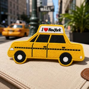 May include: An embroidered patch shaped like a yellow New York City taxi cab. The roof sign features the text I heart New York with a red heart symbol. The design includes black windows, a black checkered stripe along the side, and black wheels.