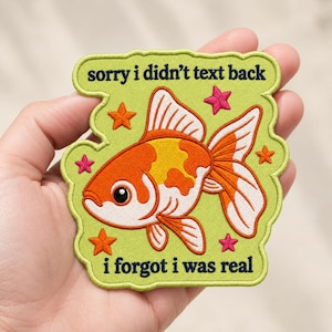 May include: A lime green embroidered patch featuring an orange and white goldfish in the center, surrounded by small pink and orange stars. The text "sorry i didn't text back" is at the top and "i forgot i was real" is at the bottom in dark blue.