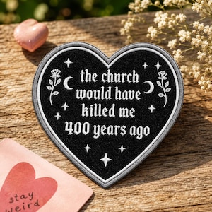 May include: A heart-shaped black embroidered patch featuring white text that reads, the church would have killed me 400 years ago. The design includes white embroidered roses, crescent moons, and small four-pointed stars surrounding the text.
