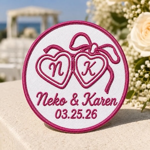 May include: A circular white embroidered patch with a dark pink border. The design features two heart-shaped lockets tied with a ribbon, containing the letters N and K. Below, the text reads Neko & Karen and the date 03.25.26 in dark pink thread.
