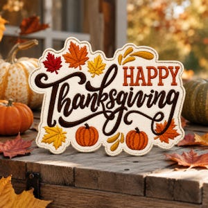 May include: An embroidered fabric sign with a cream-colored background featuring the text "HAPPY Thanksgiving" in dark brown and orange thread. The design is decorated with colorful red, orange, and yellow maple leaves and two small orange pumpkins.