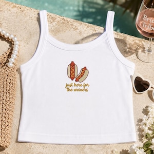 May include: A white sleeveless tank top featuring a graphic of two hot dogs and the text "just here for the weiners" printed in brown. The garment has a scoop neckline and a straight hemline, constructed with a ribbed trim along the collar and armholes.