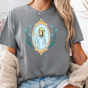 May include: Grey t-shirt featuring a cartoon illustration of a woman in a mirror, surrounded by blue flowers. The mirror has a gold frame. The word "GREY" is printed in the upper left corner.