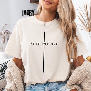 May include: Ivory-colored t-shirt with the text "FAITH OVER FEAR" and a vertical line design. The shirt is a neutral color and has short sleeves. The word "IVORY" is printed at the top of the shirt.
