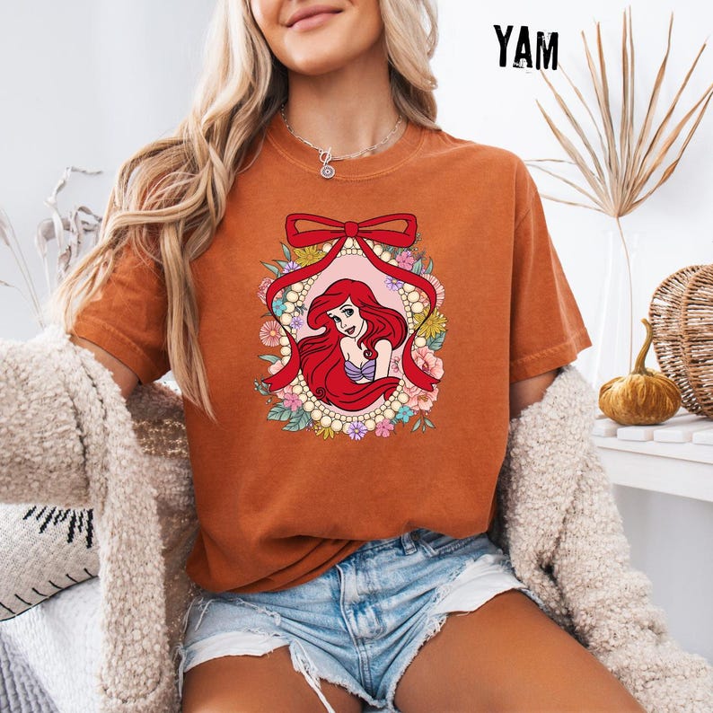 May include: Burnt orange t-shirt featuring a graphic of a red-haired cartoon character within a floral and ribbon frame. The character has a purple top and is surrounded by flowers and pearls. The word "YAM" is printed in black in the upper right corner.