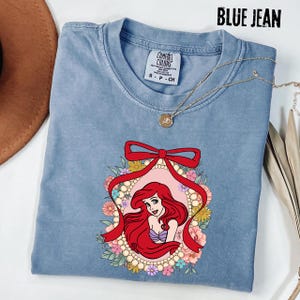 May include: A blue jean Comfort Colors t-shirt featuring a graphic of a red-haired cartoon character within a floral frame and a red bow. The shirt has a crew neck and short sleeves. A gold necklace with a pendant is draped on the shirt.