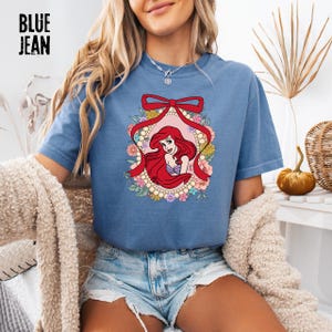 May include: Blue jean-colored t-shirt featuring a graphic of a red-haired cartoon character within a floral and ribbon frame. The text "BLUE JEAN" is printed in black at the top left of the shirt. The design includes pink, yellow, and green floral elements.