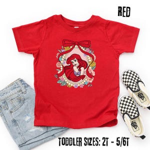 May include: A red toddler t-shirt featuring a cartoon illustration of a princess with long red hair, framed by a floral wreath and a bow. The text "RED" is at the top right. The bottom of the shirt reads "TODDLER SIZES: 2T - 5/6T".