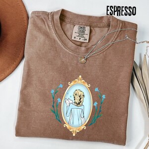 May include: Espresso-colored t-shirt with a crew neck and a graphic of a woman in a gold-framed mirror, surrounded by blue flowers. The shirt has a Comfort Colors tag and a silver necklace. The word "ESPRESSO" is printed at the top.