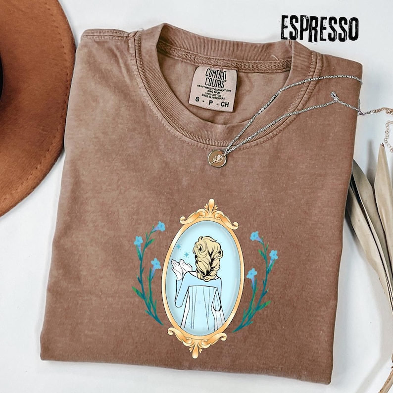 May include: Espresso-colored t-shirt with a crew neck and a graphic of a woman in a gold-framed mirror, surrounded by blue flowers. The shirt has a Comfort Colors tag and a silver necklace. The word "ESPRESSO" is printed at the top.