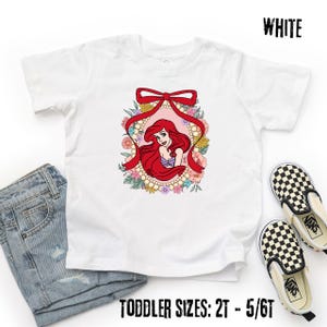 May include: White toddler t-shirt featuring a colorful graphic of a cartoon character with long red hair, framed by a red bow and floral accents. The text "WHITE" is at the top right, and "TODDLER SIZES: 2T - 5/6T" is at the bottom.