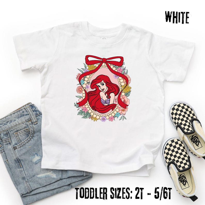 May include: White toddler t-shirt featuring a colorful graphic of a cartoon character with long red hair, framed by a red bow and floral accents. The text "WHITE" is at the top right, and "TODDLER SIZES: 2T - 5/6T" is at the bottom.