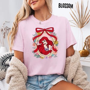May include: A light pink t-shirt featuring a cartoon illustration of a red-haired mermaid within a floral and pearl-adorned oval frame, topped with a red bow. The word "BLOSSOM" is printed in black at the top right.