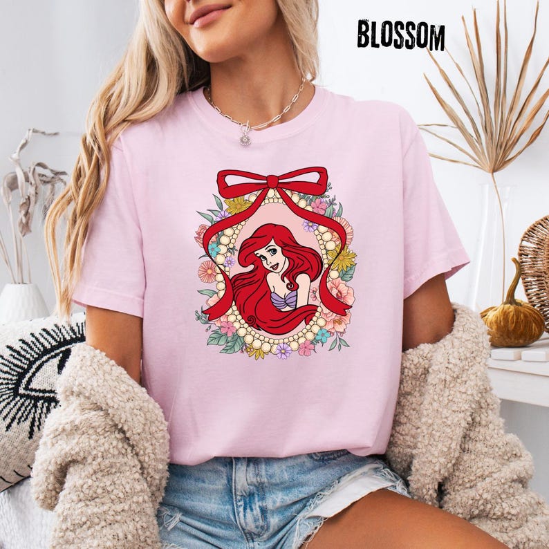 May include: A light pink t-shirt featuring a cartoon illustration of a red-haired mermaid within a floral and pearl-adorned oval frame, topped with a red bow. The word "BLOSSOM" is printed in black at the top right.