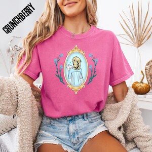 May include: A pink t-shirt with a graphic of a woman in a gold framed mirror, surrounded by blue flowers. The text "CRUNCHBERRY" is printed in black on the top left of the shirt. The shirt is a short-sleeved, crew neck style.