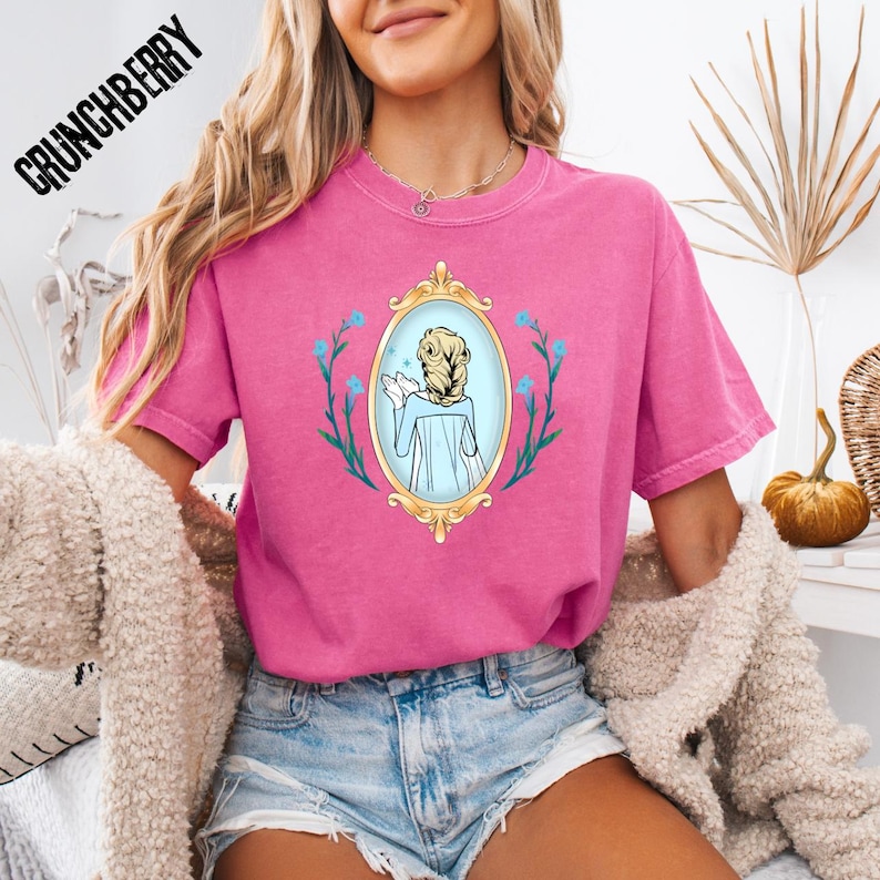 May include: A pink t-shirt with a graphic of a woman in a gold framed mirror, surrounded by blue flowers. The text "CRUNCHBERRY" is printed in black on the top left of the shirt. The shirt is a short-sleeved, crew neck style.