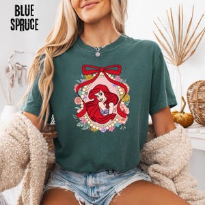 May include: Teal-colored t-shirt featuring a cartoon illustration of a red-haired mermaid within a floral and pearl-adorned frame, accented by a red bow. The words "BLUE SPRUCE" are printed in black at the top left.