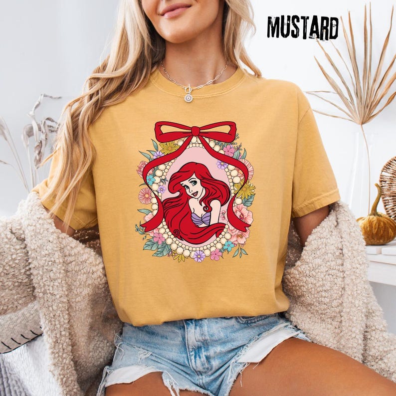 May include: Mustard-colored t-shirt featuring a graphic of a red-haired mermaid within a floral and pearl-adorned frame, accented by a red ribbon. The word "MUSTARD" is printed in black at the top right.