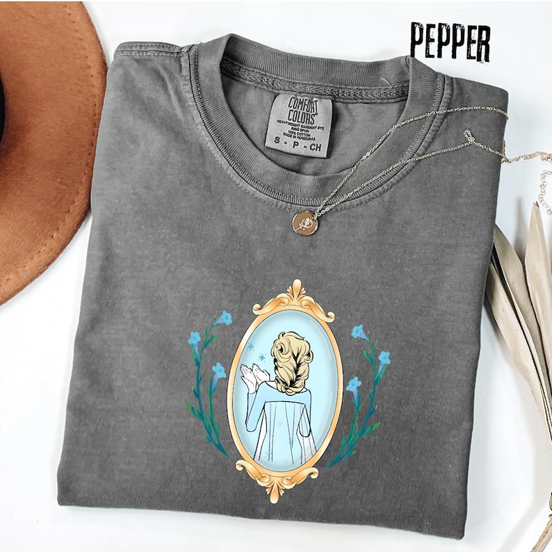 May include: A gray Comfort Colors t-shirt featuring a gold-framed oval mirror design with a cartoon illustration of a woman with blonde hair and a blue dress. The shirt has a crew neck and short sleeves. A silver necklace with a pendant is draped across the shirt.