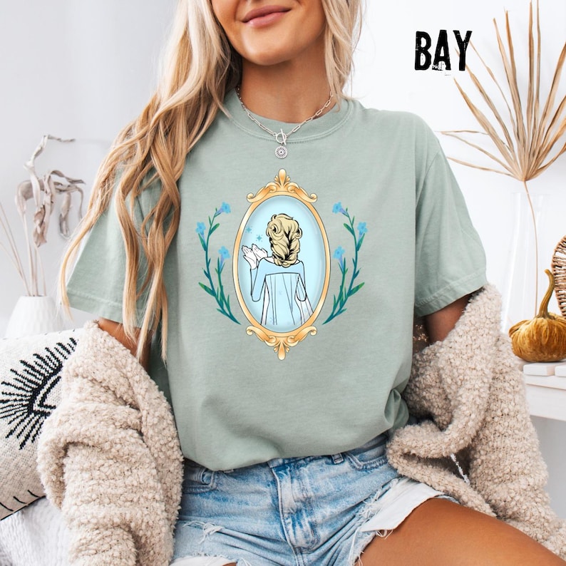 May include: Sage green t-shirt featuring a gold framed mirror graphic with a blonde woman's reflection. The mirror is surrounded by blue flowers and green vines. The word "BAY" is printed in black at the top right.