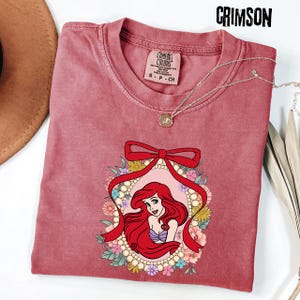 May include: Crimson-colored t-shirt featuring a graphic of a red-haired woman within a floral frame and a red bow. The shirt has a crew neck and short sleeves. A silver necklace with a pendant is draped across the shirt.