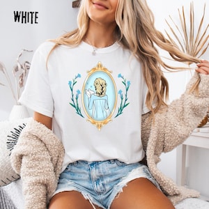 May include: White t-shirt with a gold framed mirror graphic. The mirror shows a woman with blonde hair in a blue dress. Blue flowers and green leaves surround the mirror. The word "WHITE" is printed in black at the top left.