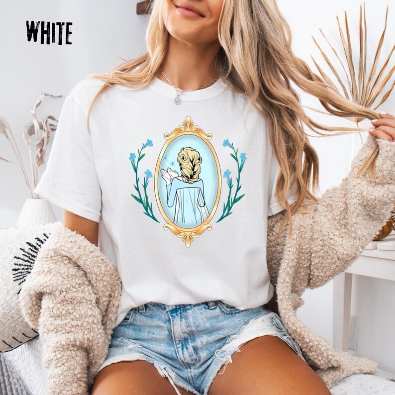 May include: White t-shirt with a gold framed mirror graphic. The mirror shows a woman with blonde hair in a blue dress. Blue flowers and green leaves surround the mirror. The word "WHITE" is printed in black at the top left.