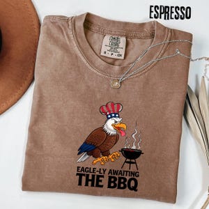 Könnte beinhalten: Espressofarbenes T-Shirt mit einem Cartoon-Adler, der einen Kochhut mit roten, weißen und blauen Akzenten trägt. Der Adler grillt und der Text lautet "EAGLE-LY AWAITING THE BBQ". Eine silberne Halskette mit Anhänger befindet sich auf dem Shirt.