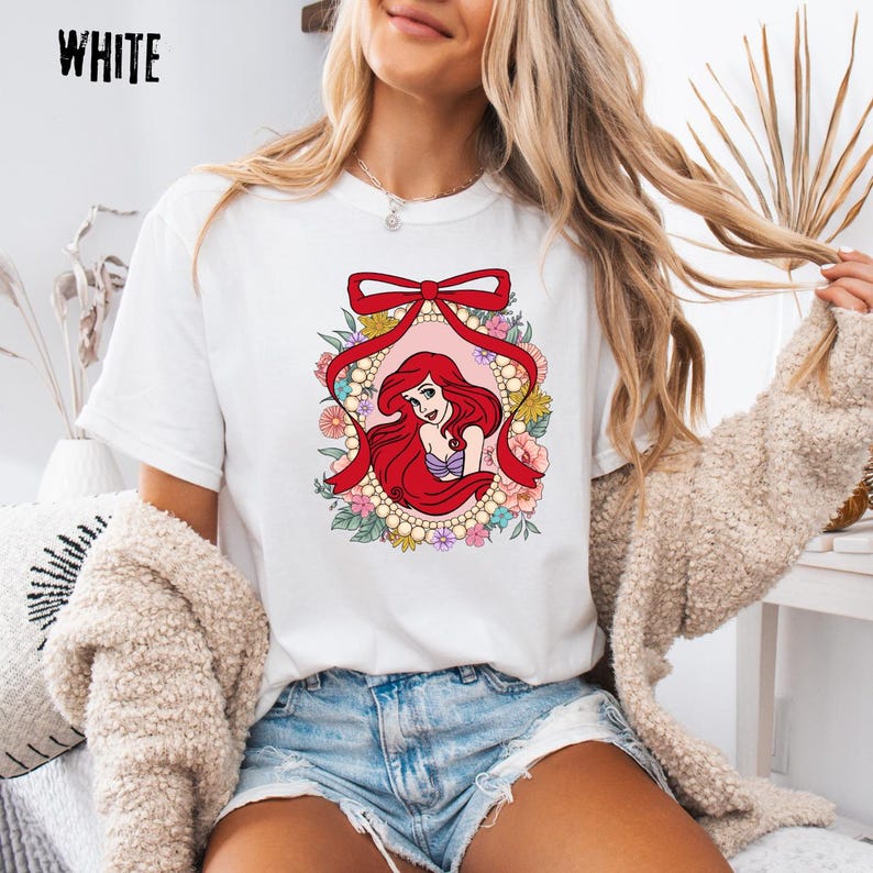 May include: White t-shirt featuring a cartoon illustration of a red-haired mermaid within a floral and pearl-framed oval, topped with a red bow. The word "WHITE" is printed in black at the top left.