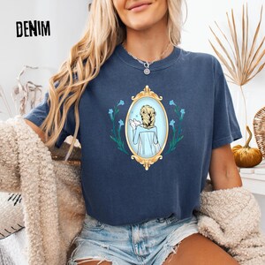 May include: A navy blue t-shirt with the word "DENIM" printed at the top. The shirt features a gold framed oval design with a light blue illustration of a person with blonde hair. The design is surrounded by blue flowers.