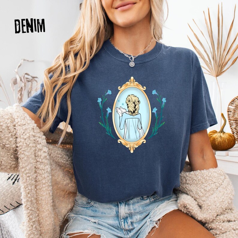 May include: A navy blue t-shirt with the word "DENIM" printed at the top. The shirt features a gold framed oval design with a light blue illustration of a person with blonde hair. The design is surrounded by blue flowers.
